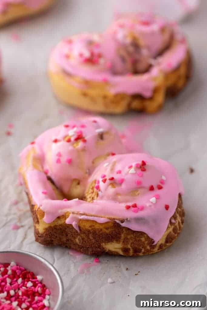 Valentines Sweetheart Cinnamon Buns Made Easy 13 Several heart-shaped cinnamon rolls, beautifully iced with pink frosting and sprinkles, arranged on a baking sheet lined with parchment paper.