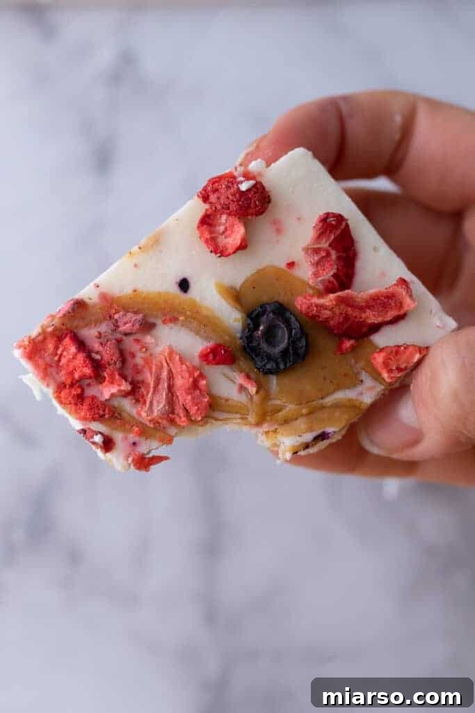 Protein Packed Cottage Cheese Freezer Bark 9 A hand holding a single square of frozen cottage cheese bark, with a bite taken out, showing its creamy interior and berry topping.