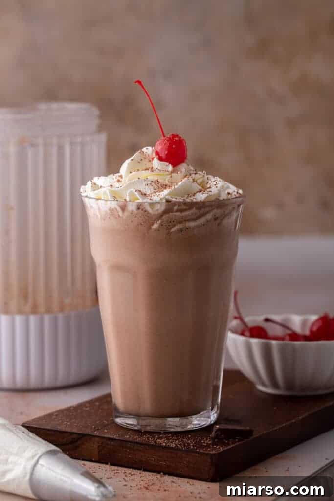A thick, creamy chocolate milkshake in a tall glass, with a blender and ingredients in the background.