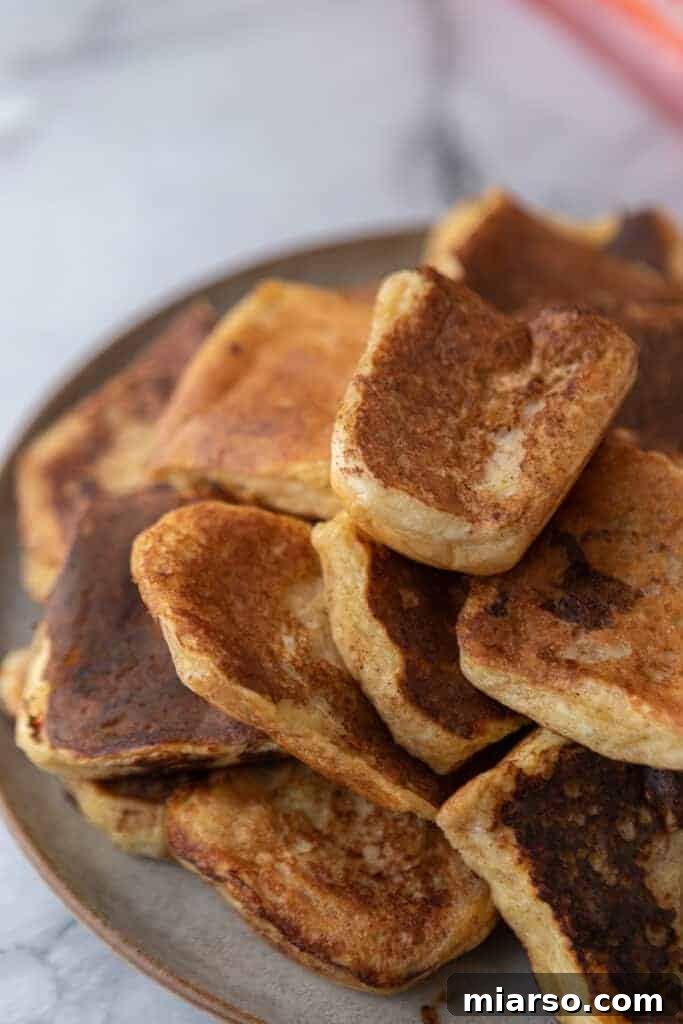 A stack of golden-brown, freshly cooked Hawaiian roll French toast pieces on a white plate, ready for toppings.