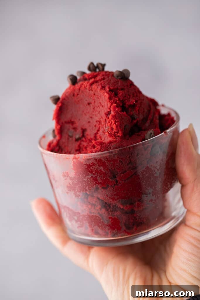 Hand holding a glass cup filled with edible red velvet cookie dough, showing its rich texture and color.