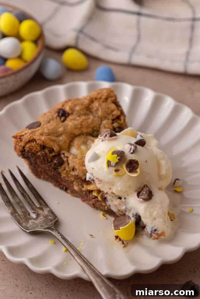 Gooey Giant Cadbury Mini Egg Skillet Cookie 4 A slice of Cadbury Mini Egg Skillet Cookie on a plate, topped with a scoop of vanilla ice cream and a fork, ready to be enjoyed.