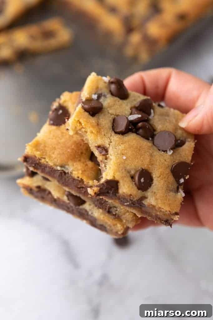 A hand gently holding a perfectly baked, warm, and gooey chocolate chip cookie bar, displaying its rich texture and inviting appearance.