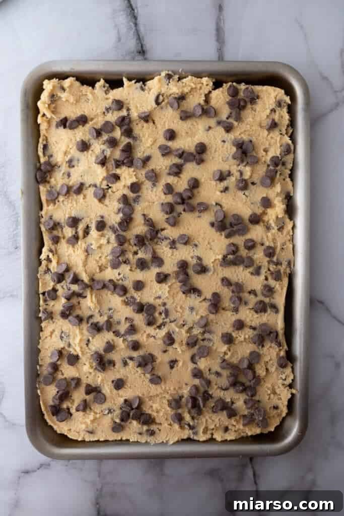 The chocolate chip cookie dough spread evenly across a baking sheet, topped with extra chocolate chips for a beautiful finish, before entering the oven.