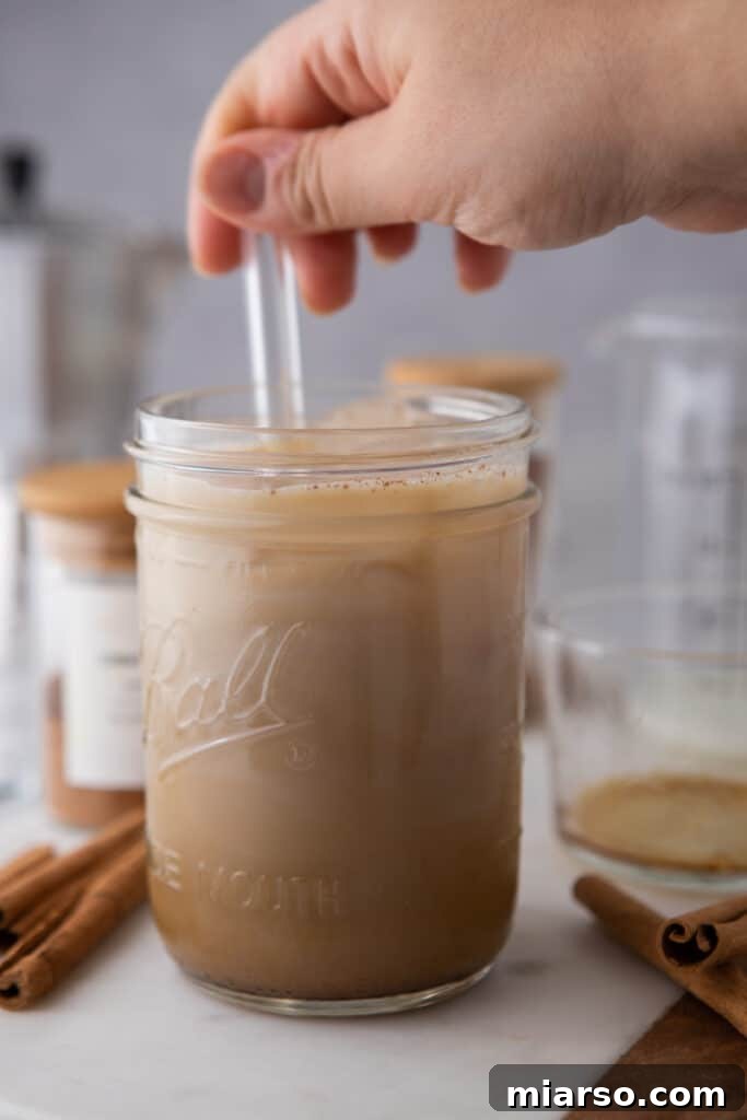 A hand stirring an iced latte with a straw in a glass, mixing the espresso, milk, and maple syrup.