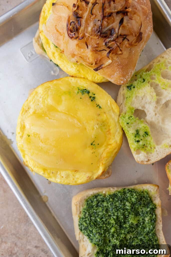 Assembled ingredients on a baking sheet, featuring toasted ciabatta with melted cheese, egg patties, pesto, and mozzarella, ready to be put together.