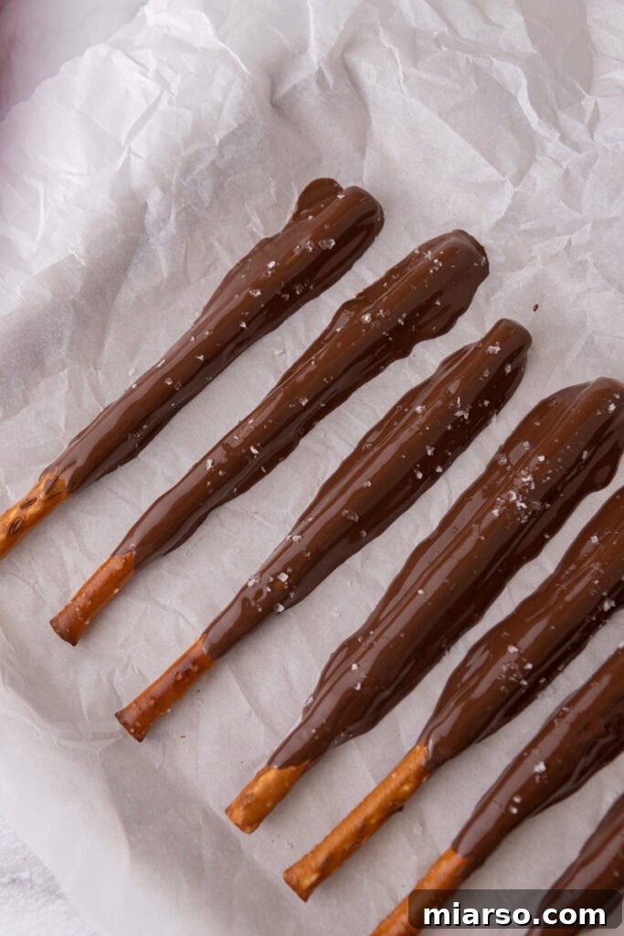 Decadent Caramel Chocolate Pretzel Rods 10 Freshly chocolate-coated pretzel rods, some sprinkled with sea salt, resting on a baking sheet as they set.