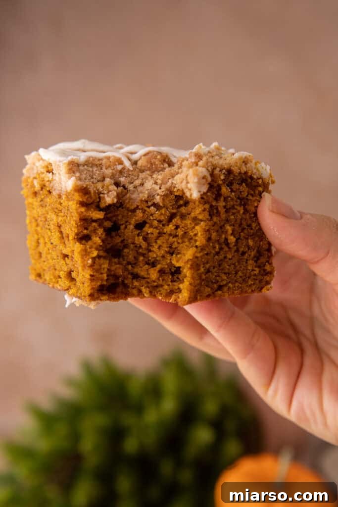 A slice of pumpkin coffee cake, featuring the moist cake, streusel, and glaze, held against a blurred autumn background.