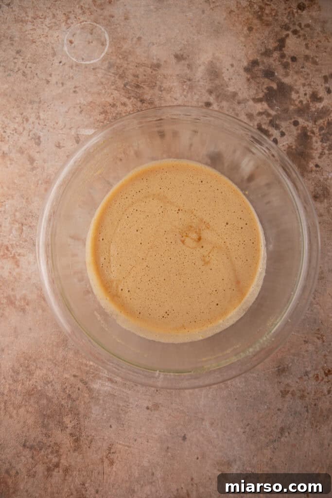 A mixing bowl containing whisked eggs, vanilla extract, and various sugars for the pecan pie filling, showing a smooth and well-combined base.