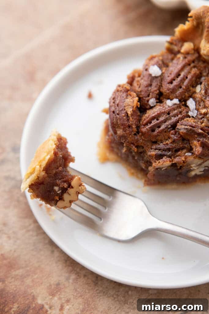 An overhead shot of a perfectly baked slice of maple pecan pie on a white plate, with a fork poised to take a bite, showcasing the rich, glistening filling and crunchy pecan topping.
