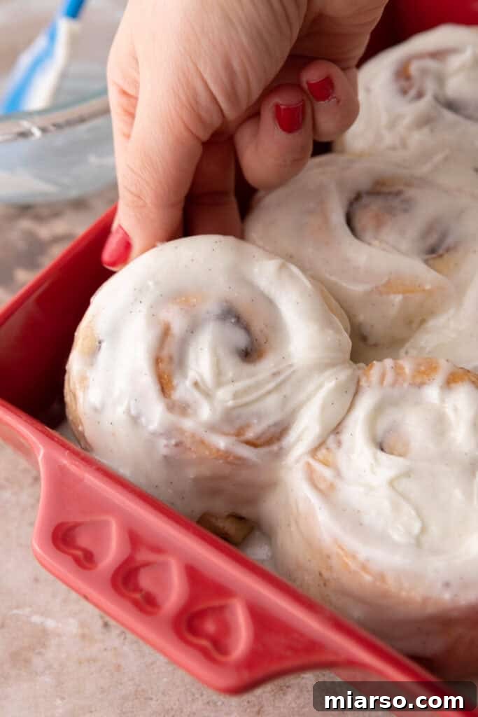 Perfect Small Batch Cinnamon Rolls 12 Hand lifting a freshly baked small-batch cinnamon roll covered in creamy vanilla bean frosting from a red baking dish.