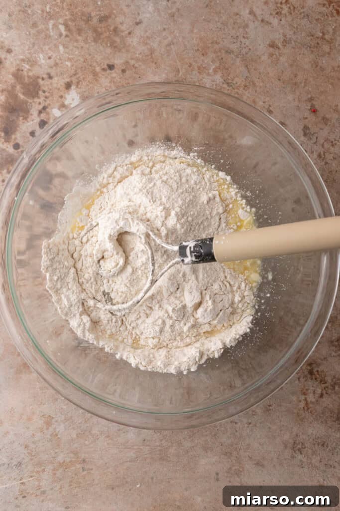 Perfect Small Batch Cinnamon Rolls 5 Flour being mixed into cinnamon roll dough ingredients in a glass bowl using a dough whisk.
