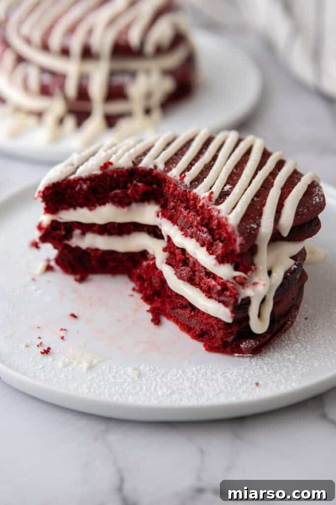 Indulgent Red Velvet Pancakes 11 A large stack of Red Velvet Pancakes on a plate, generously covered with a thick layer of cream cheese frosting and a drizzle, with powdered sugar.