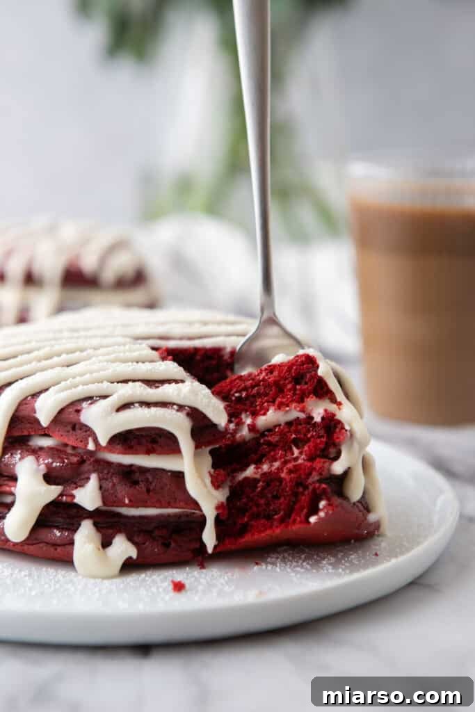 Indulgent Red Velvet Pancakes 4 A stack of freshly cooked Red Velvet Pancakes Recipe on a white plate, drizzled with cream cheese glaze and dusted with powdered sugar, ready to be enjoyed.