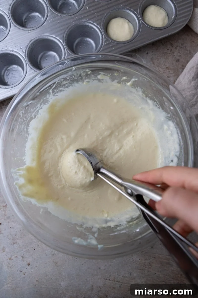 Scooping the thick, elastic Brazilian cheese bread batter into the greased cavities of a mini muffin pan, filling each about halfway.