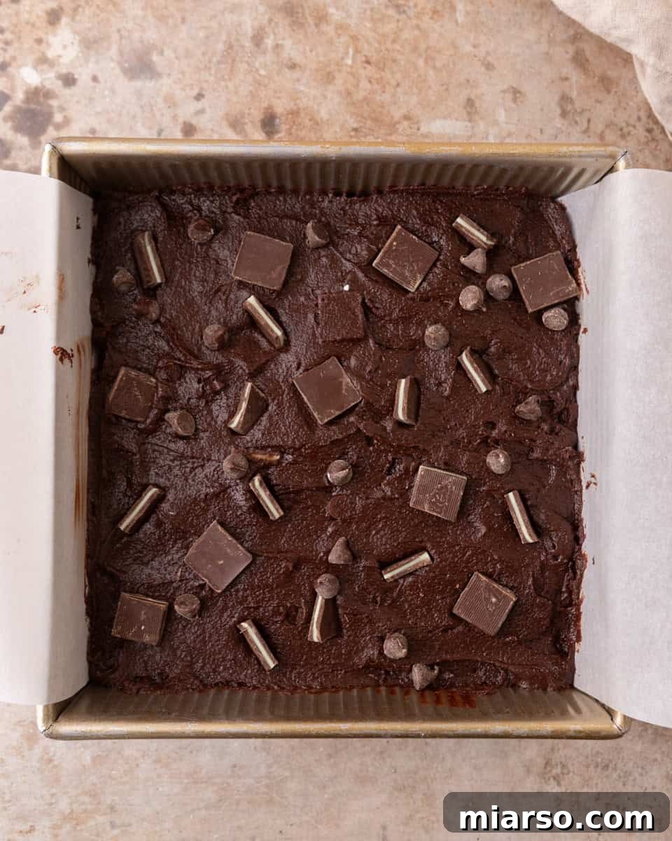 Ultra Fudgy Crinkled Andes Mint Brownies 9 Brownie batter spread evenly in a lined baking pan, topped with chocolate and mint pieces, ready for the oven.