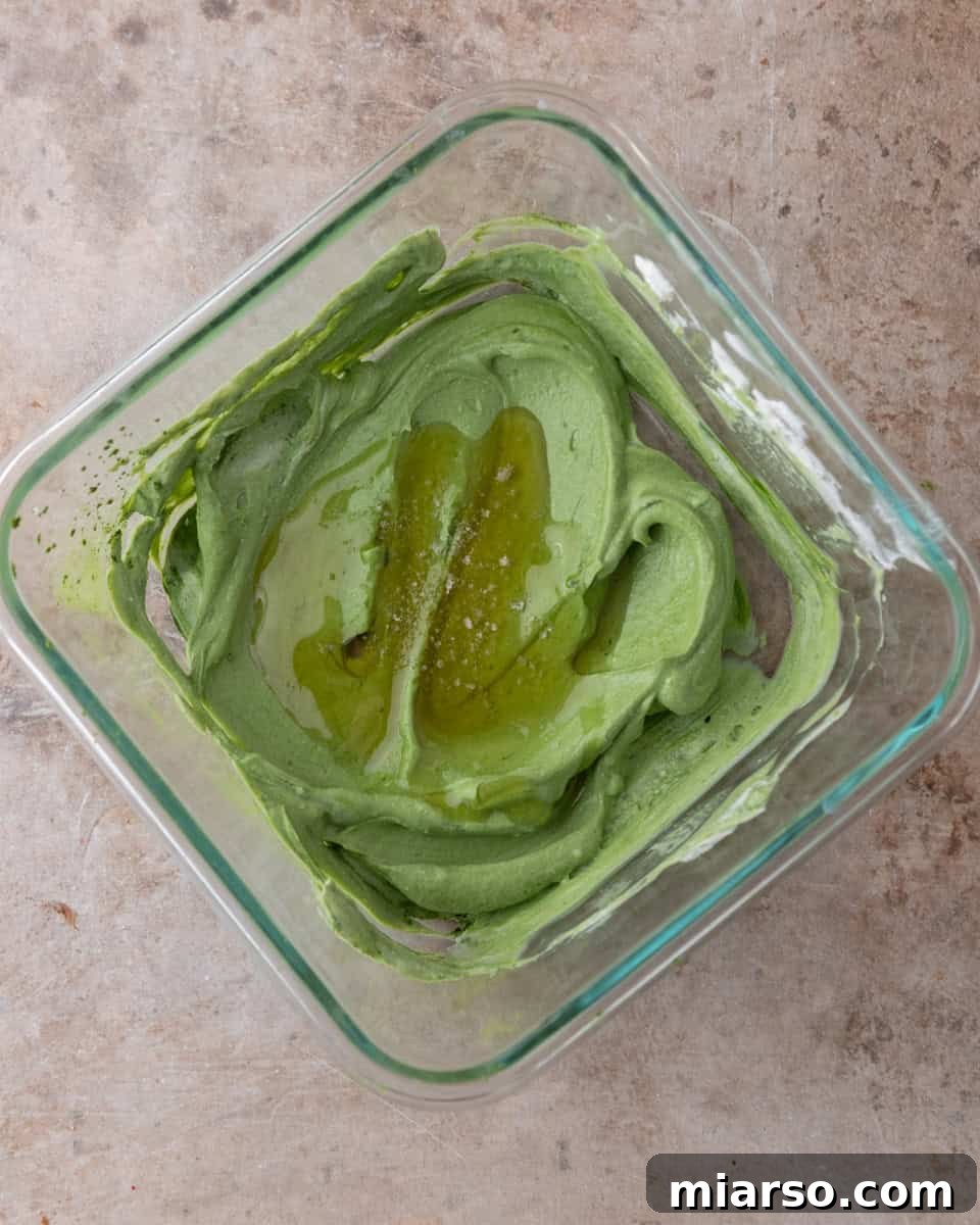 A bowl containing a smooth, vibrant green matcha yogurt mixture, with a spoon having just stirred in maple syrup and salt, ready for milk addition.