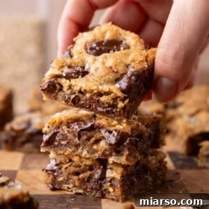 A hand lifting a chewy brown butter cookie bar from a stack on a wooden cutting board, showing melted chocolate and flaky sea salt on top
