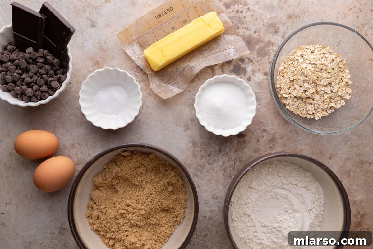 Ingredients for cookie bars laid out in bowls including butter, brown sugar, flour, rolled oats, eggs, chocolate chips, and chocolate chunks