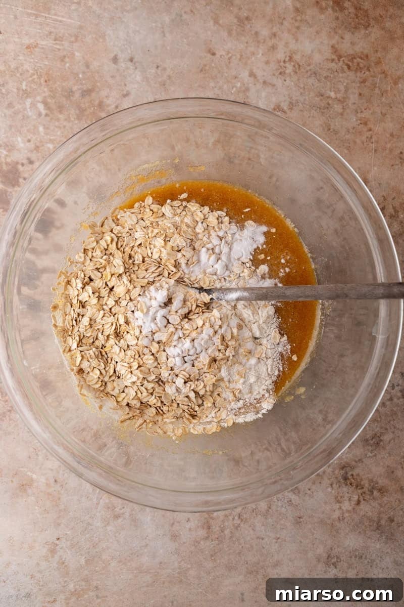 Rolled oats, flour, and baking soda being stirred into wet batter with a spoon in a glass bowl
