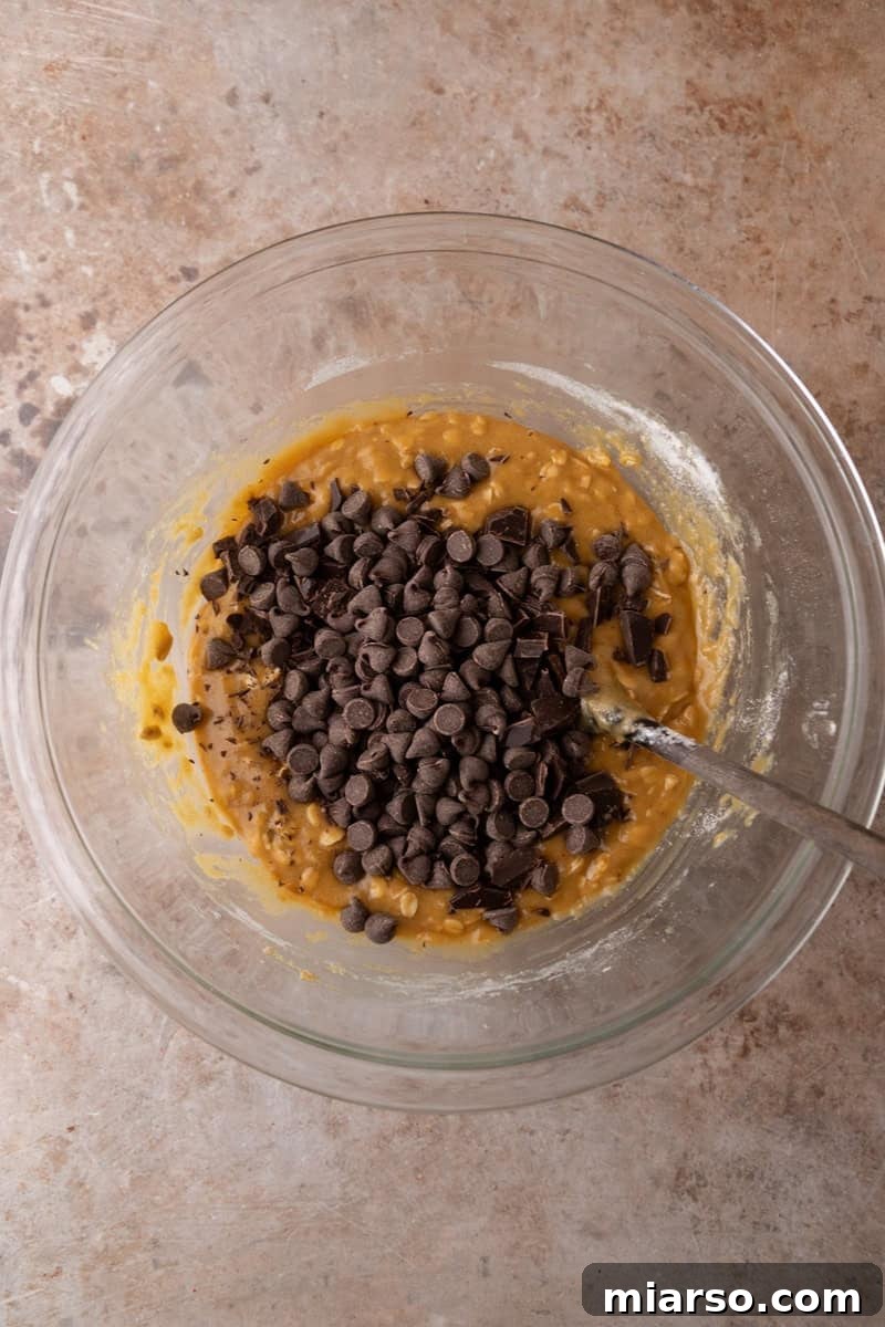 Chocolate chips and chocolate chunks poured on top of cookie bar dough in a glass mixing bowl