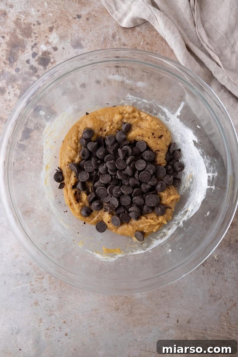Nutty Brown Butter Chocolate Chip Skillet Cookie 6 Thick cookie dough in a glass bowl with dark chocolate chips piled on top before folding them in