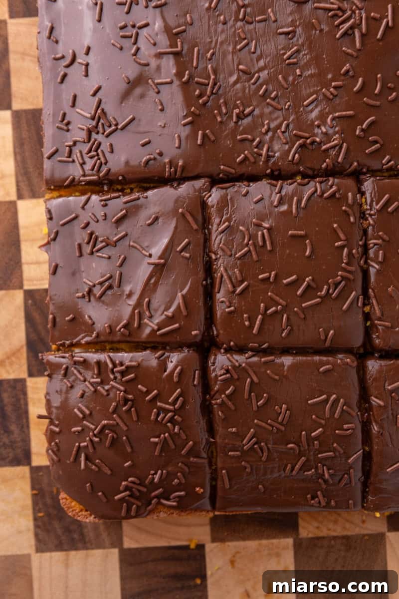 Overhead shot of chocolate-glazed cake sliced into squares on a checkered wood board with chocolate sprinkles on top
