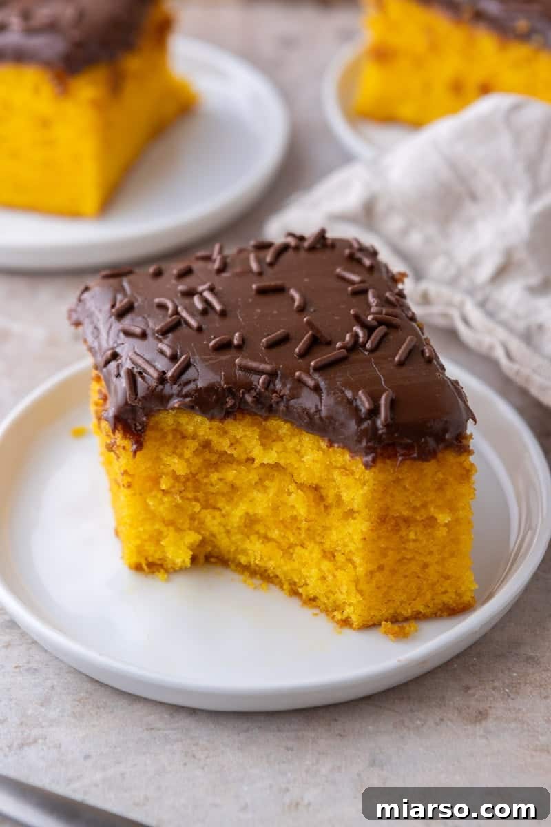 A square slice of Brazilian carrot cake with chocolate glaze on a white plate with more slices in the background