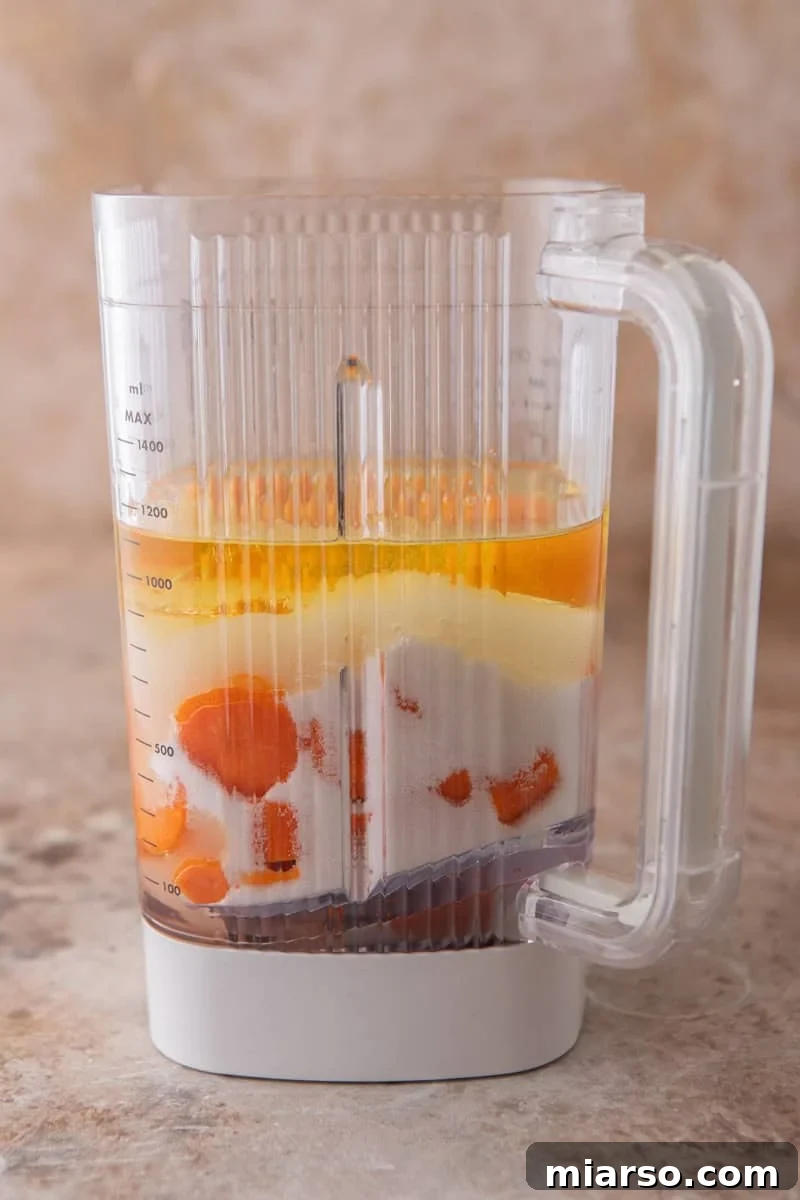 A blender filled with layers of chopped carrots, sugar, oil, and eggs before blending