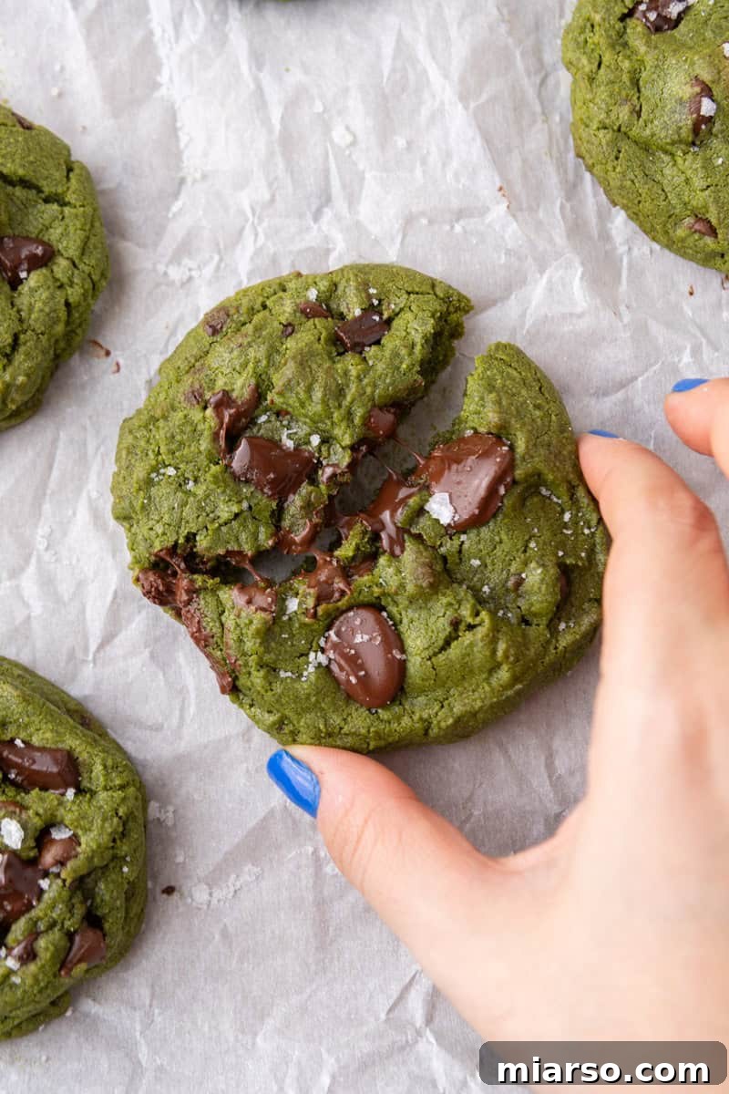 Velvet Matcha Chocolate Chip Cookies 4 Hand breaking apart a thick chewy matcha cookie with melted chocolate and sea salt flakes on parchment paper