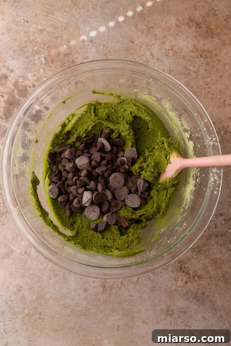 Velvet Matcha Chocolate Chip Cookies 9 Bright green cookie dough with semi-sweet chocolate chips being folded in with a pink spatula