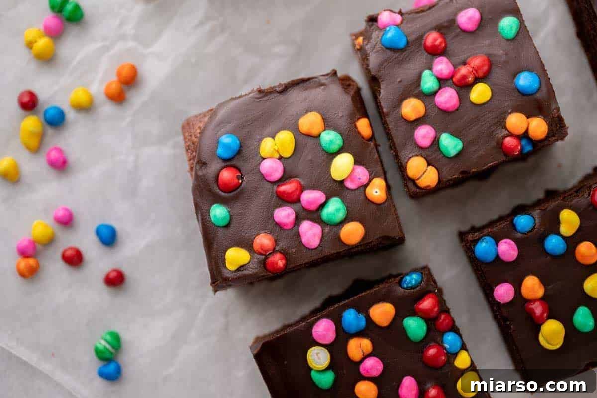 Homemade cosmic brownies, lavishly topped with a thick chocolate ganache and vibrant colorful rainbow candy chips, arranged on parchment paper.