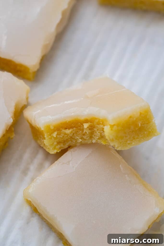 Vibrant lemon blondies with a smooth, glossy lemon glaze, neatly sliced into squares, revealing their bright yellow, fudgy interior, with one piece playfully showing a bite taken out.