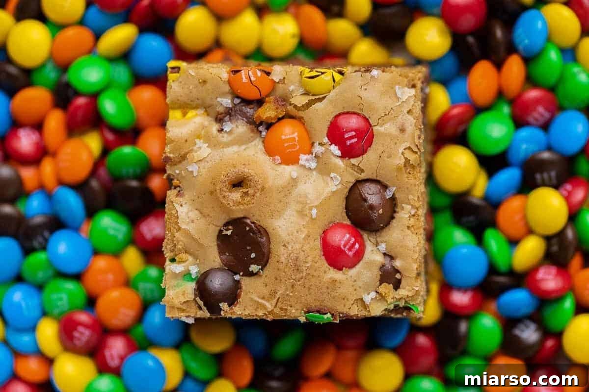A single, delightful M&M chocolate chip blondie bar, surrounded by a scatter of colorful M&M candies, showcasing its perfectly chewy texture and embedded candy pieces.