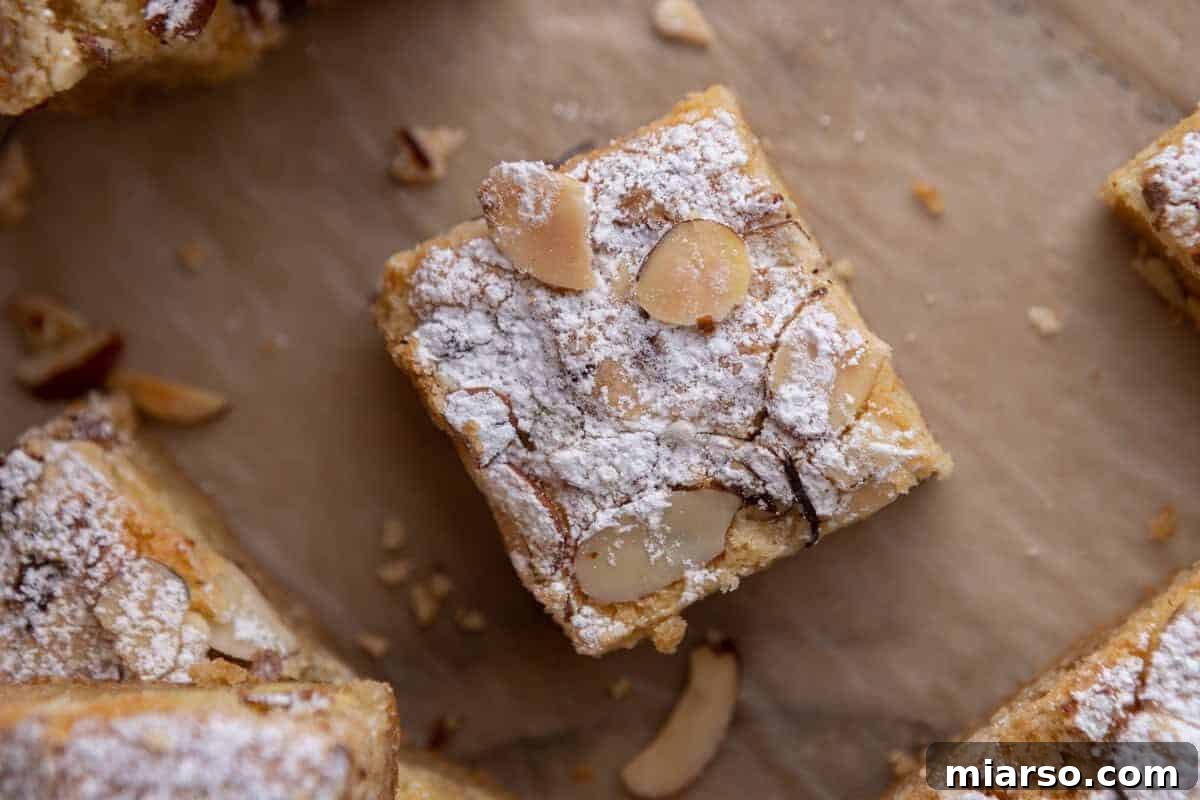 A sophisticated almond croissant blondie bar, elegantly topped with sliced almonds and a dusting of powdered sugar on parchment paper, showcasing its rich frangipane filling.