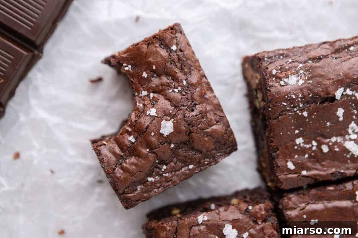 Overhead view of fudgy homemade brownies with glistening crinkly tops and delicate flaky sea salt, arranged on parchment paper beside gourmet dark chocolate squares.