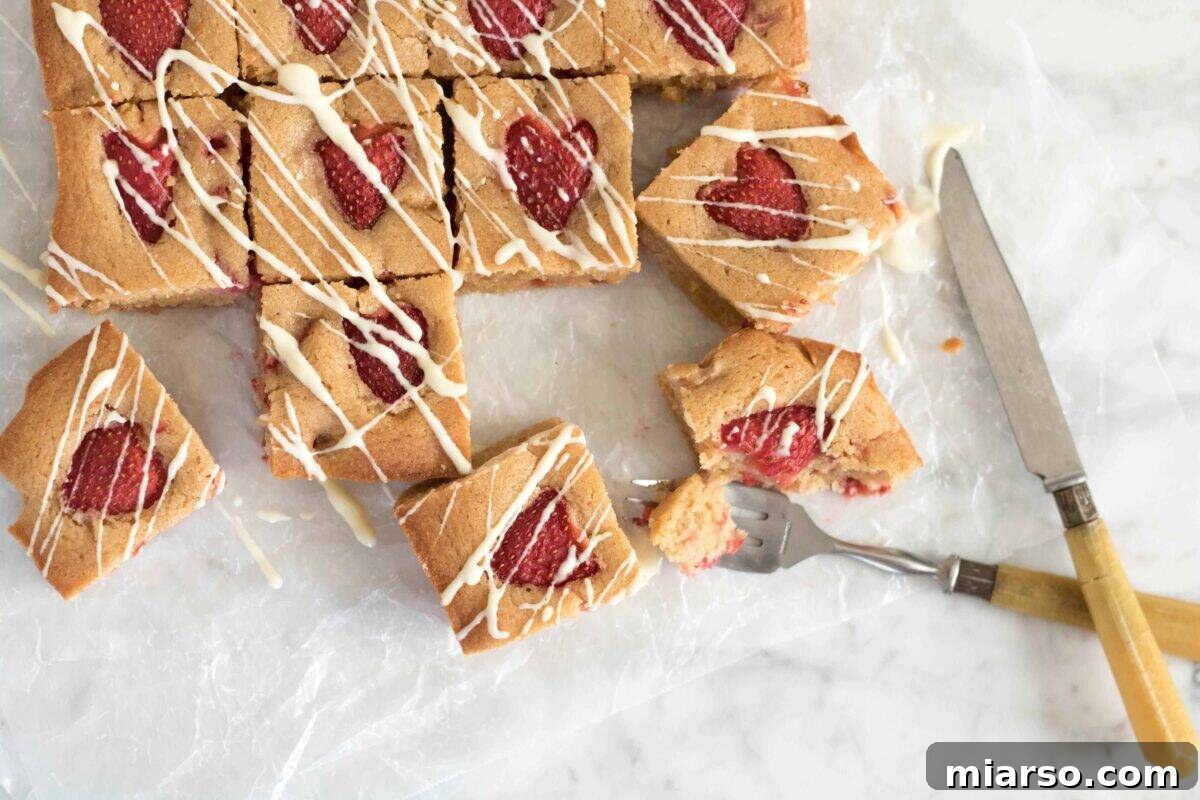 Vibrant brown butter strawberry blondies, delicately drizzled with white chocolate on parchment paper, with fresh strawberry halves beautifully baked into each square.