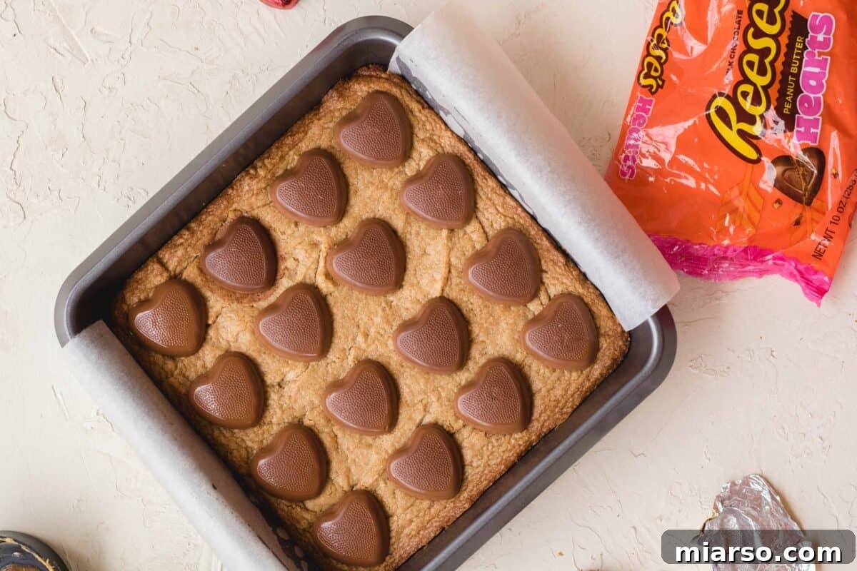 Rich peanut butter blondies baking in a pan, generously topped with festive Reese's peanut butter hearts, with the candy bag subtly visible nearby.
