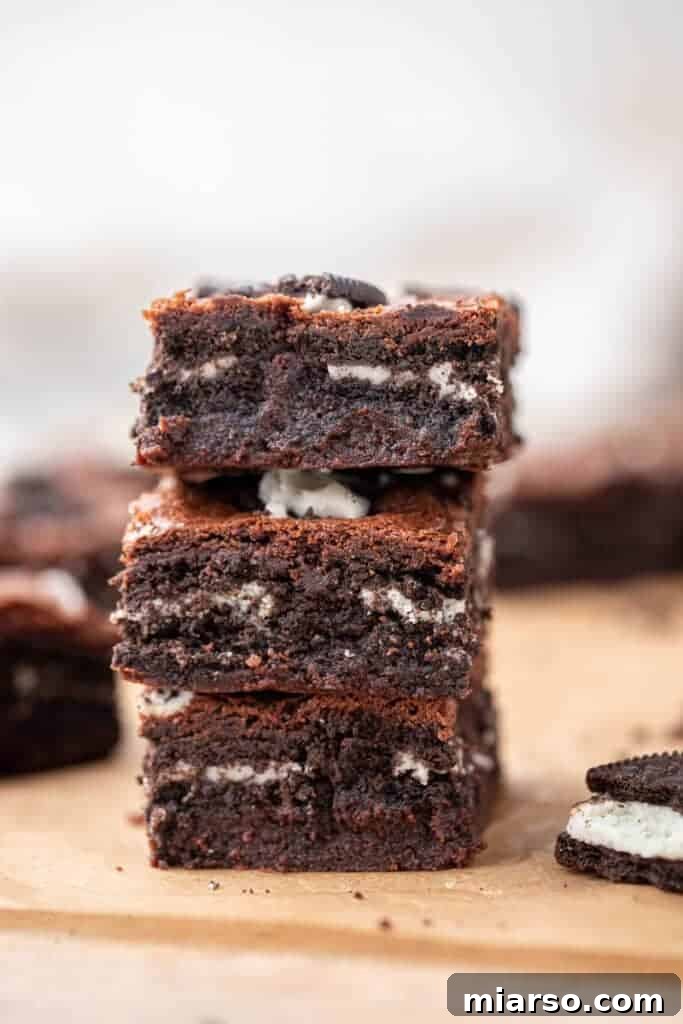 A towering stack of three fudgy Oreo brownies, revealing decadent layers of Oreo cookies baked directly into the dense chocolate interior, with extra Oreo pieces scattered on top.
