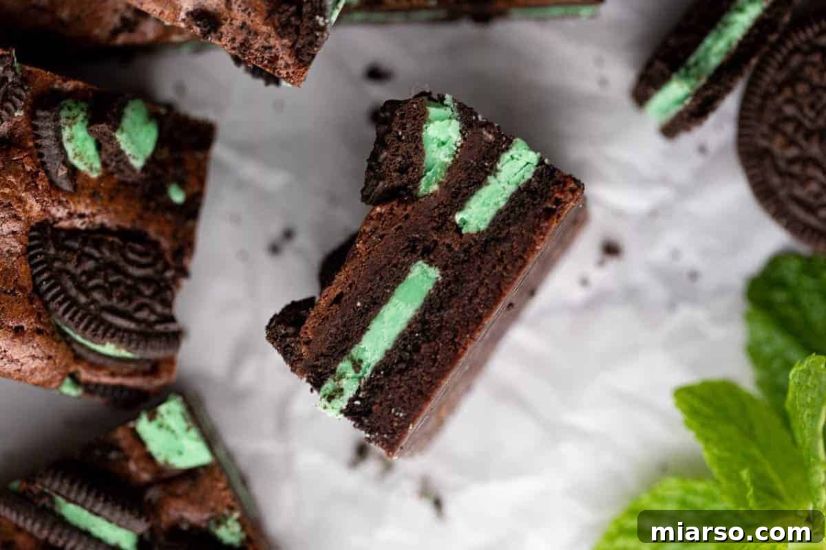 A luscious Mint Oreo brownie, artfully tilted to reveal its cross-section with vibrant layers of bright green mint Oreo filling baked into the fudgy chocolate brownie, elegantly surrounded by fresh mint leaves and mint Oreo cookies.