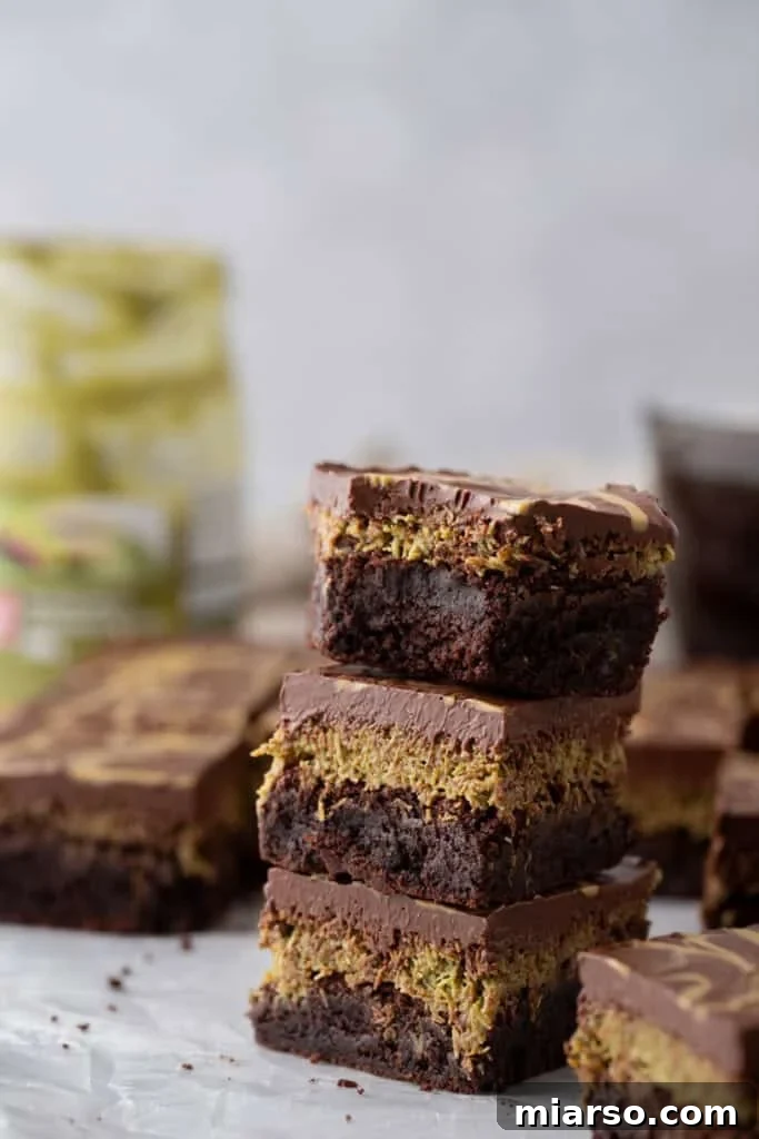 A decadent stack of three Dubai chocolate brownies, showcasing an intricate pistachio kunafa layer nestled between a rich fudgy brownie base and a thick, glossy chocolate ganache topping.