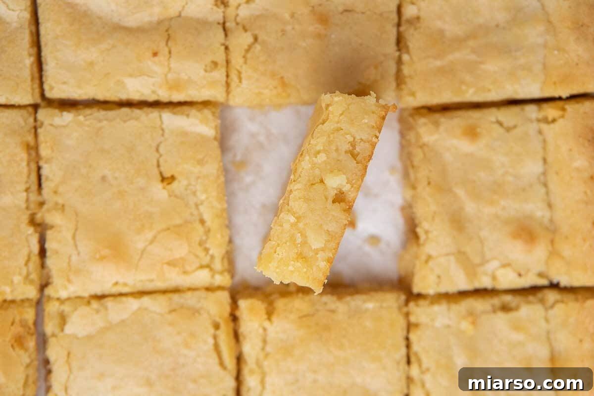 Glistening white chocolate brownies, perfectly sliced into squares, with one piece tilted to showcase its dense, fudgy golden interior and distinct crinkly top.