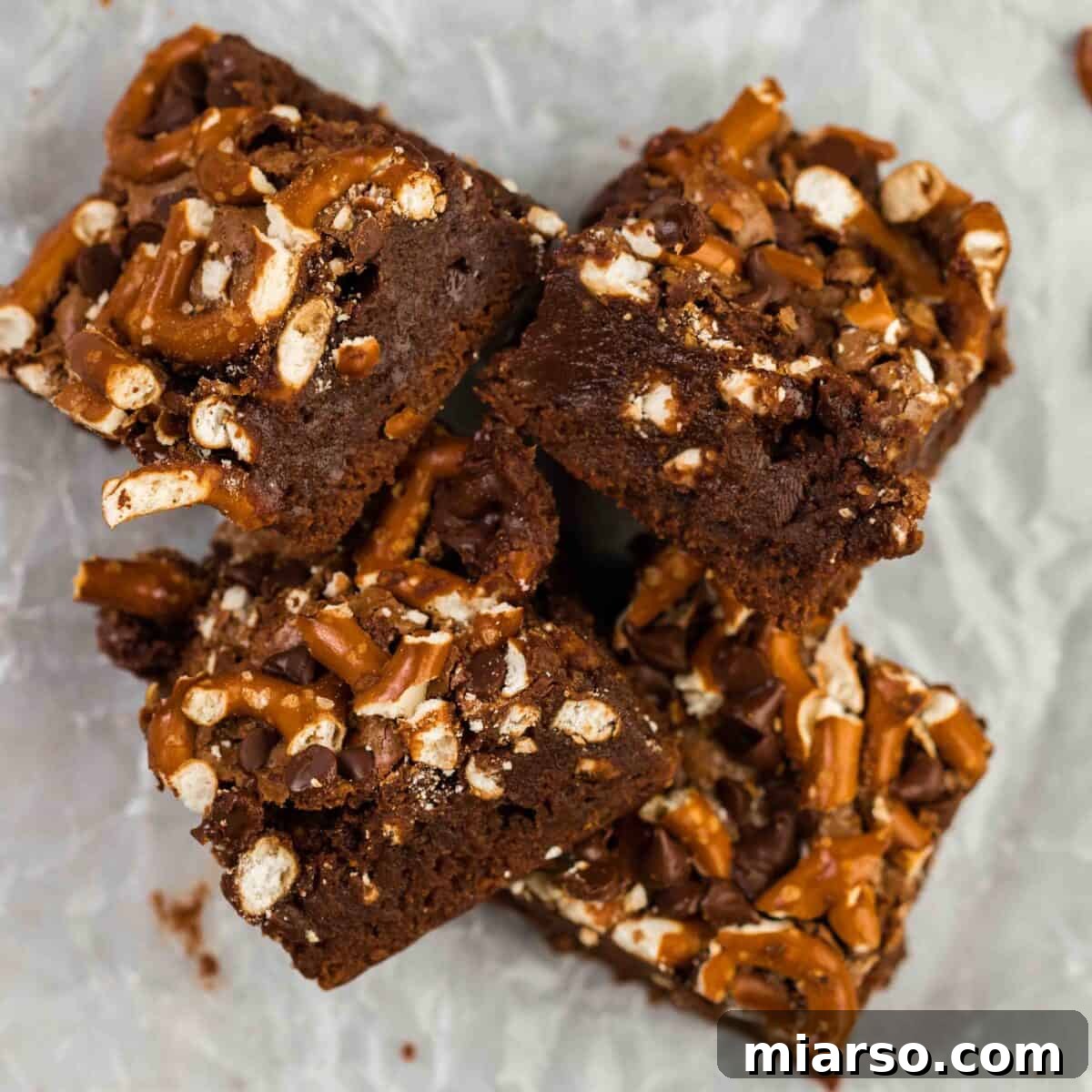 Irresistible pretzel brownies, artfully piled together, showcasing crunchy crushed pretzels and melty chocolate chips baked into their fudgy chocolate brownie base on parchment paper.