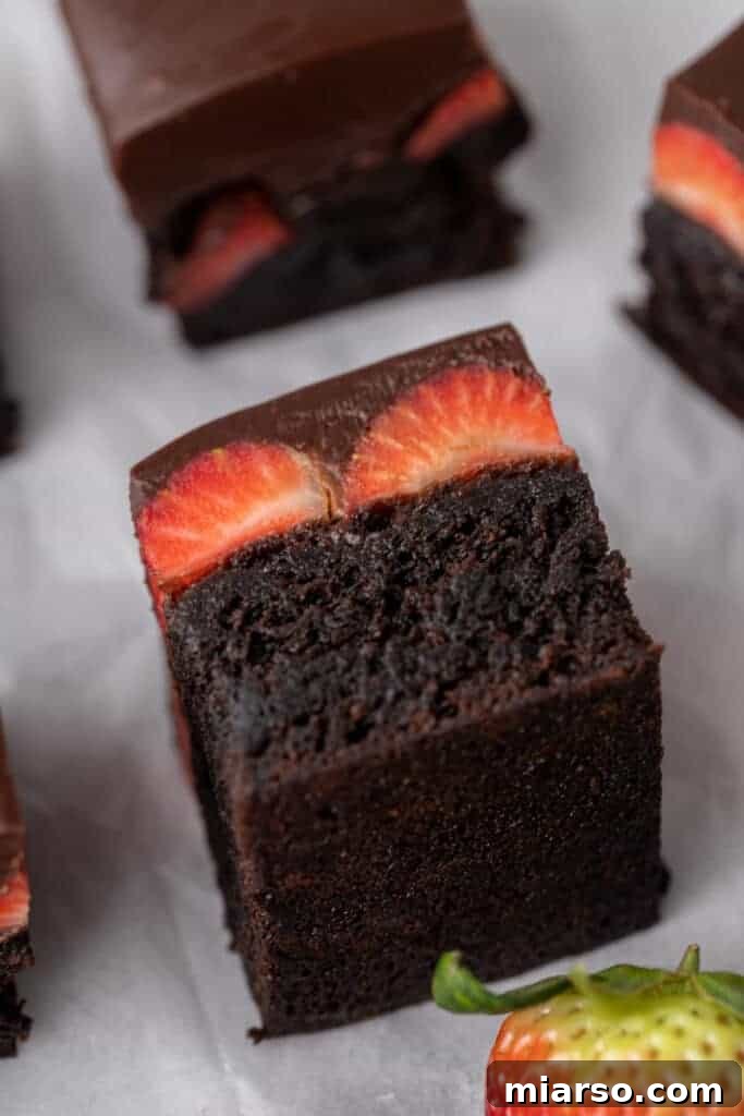 A rich chocolate-covered strawberry brownie square, showcasing a thick fudgy brownie base generously topped with glossy chocolate ganache and fresh strawberry slices.