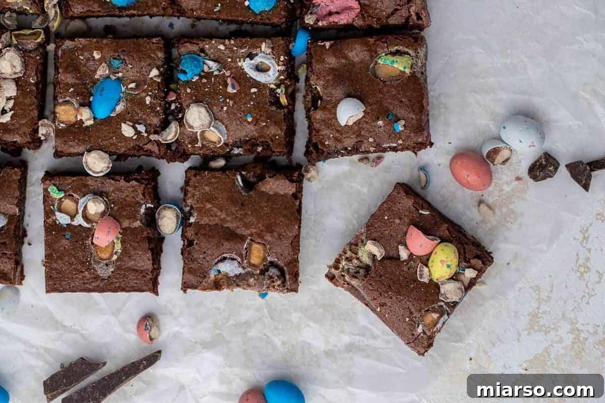 Overhead view of delectable fudgy Easter mini egg brownies, perfectly sliced into squares on parchment paper, generously topped with crushed colorful Cadbury Mini Eggs and Robin Eggs.