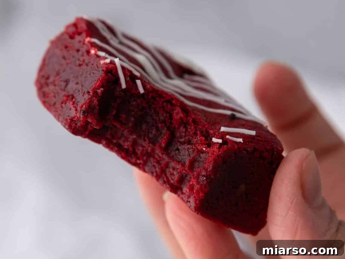 A hand holds a luscious fudgy red velvet brownie with a bite taken out, revealing its dense, vibrant red interior and elegant white chocolate drizzle on top.
