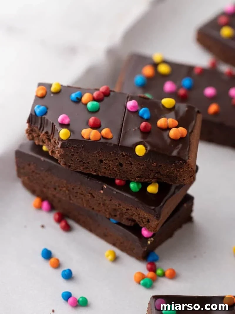 A decadent stack of three homemade cosmic brownies, featuring a thick chocolate ganache and vibrant colorful rainbow candy chips on top, with extra candies scattered around for a playful touch.