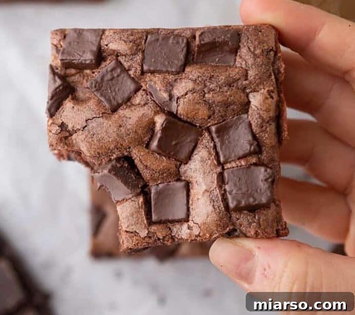 A hand holds a tempting Starbucks copycat double chocolate brownie, generously loaded with chocolate chunks on top, with a warm cup of coffee and more brownies subtly in the background.