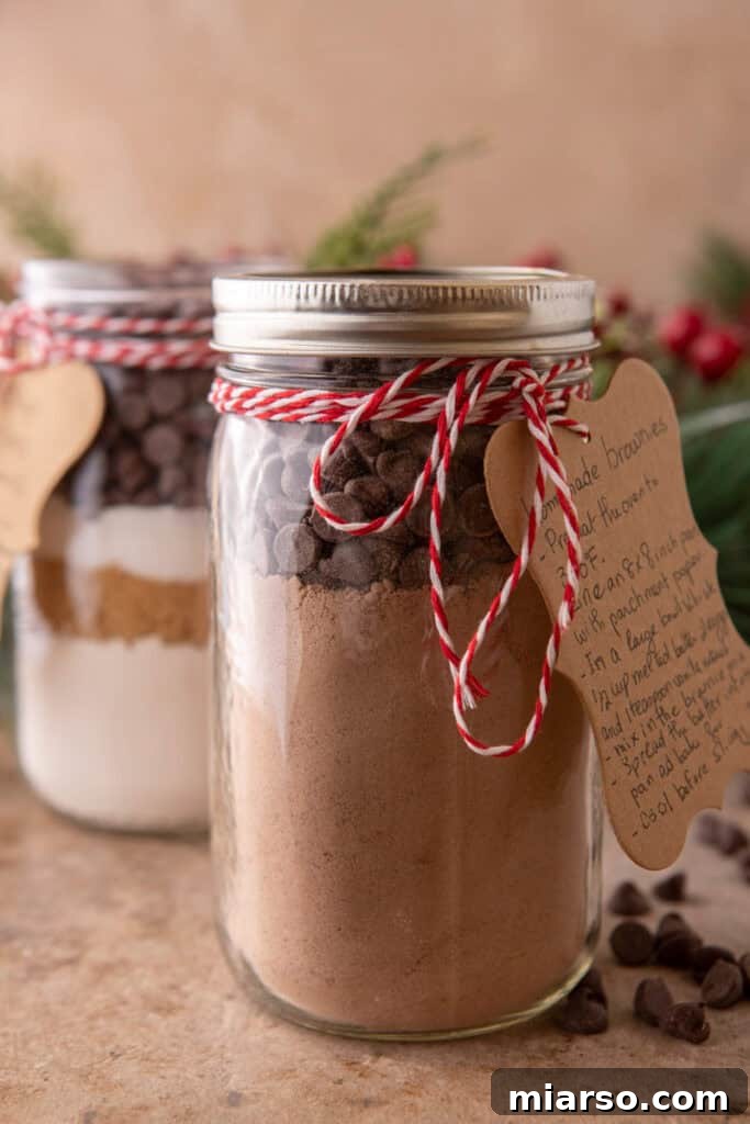 Two charming mason jars, meticulously filled with layered homemade brownie mix dry ingredients, tied with festive red and white baker's twine and adorned with handwritten recipe tags, all set against a backdrop of delicate holiday greenery.