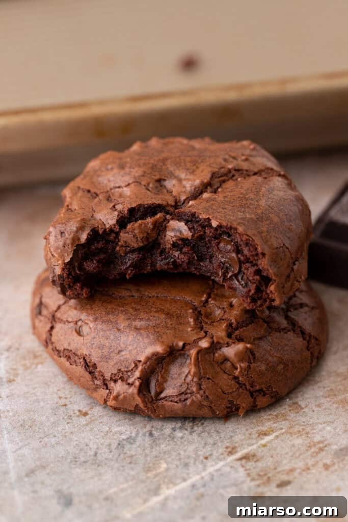 Two small batch brownie cookies, elegantly stacked, with the top cookie broken in half to reveal its irresistibly fudgy, melted chocolate interior, alongside dark chocolate squares.
